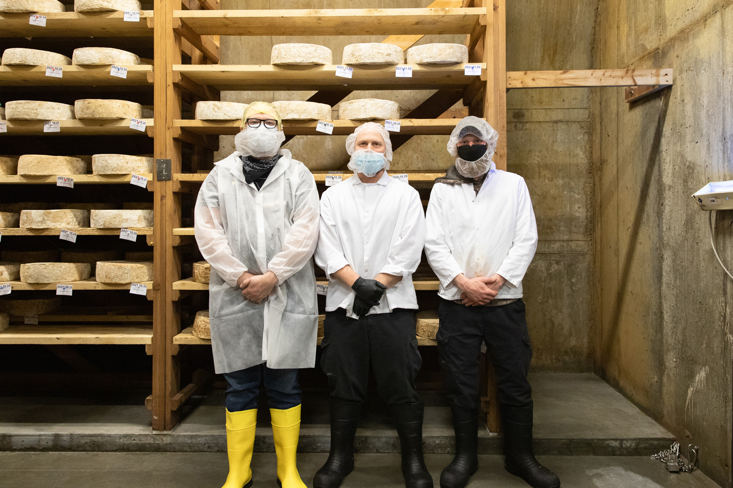 Three men stand in a cellar in front of a large shelf of cheese aging 