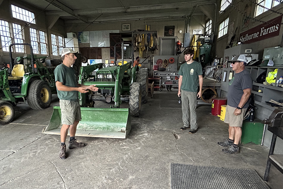 three men inside barn full of tools and tractors discuss the day's schedule