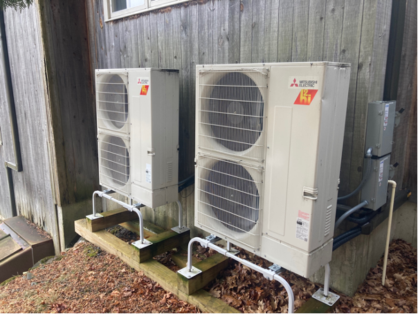 Exterior units of two air-source heat pump against wooden-sided wall of a house