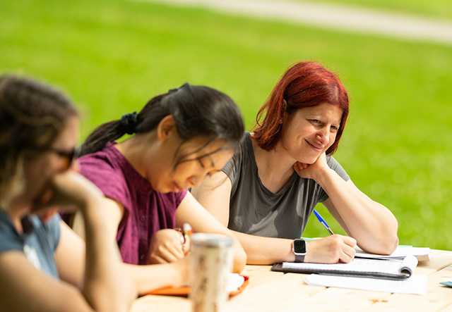 Three women sit around an outdoor picnic table, smiling as they take notes