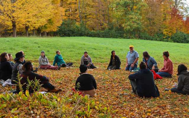 Sixteen adults sit in a circle outside on a grassy lawn in discussion with fall foliage in the background