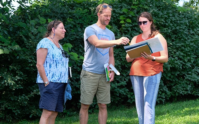 Three people stand looking at a clipboard outside, standing in front of a green hedge
