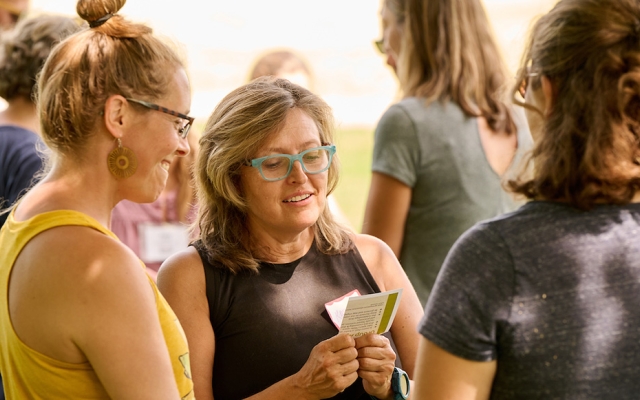 Three women stand in a circle outside in summertime looking at small flashcards