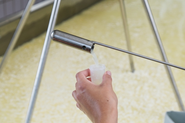 A hand holds a sample vile as a narrow metal sampling ladle pours a semi-transparent liquid into the vile.