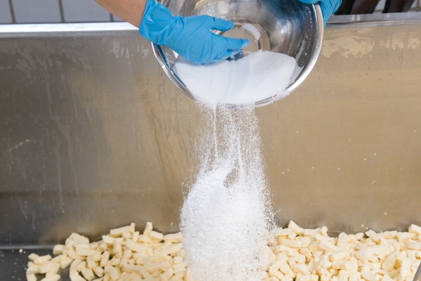 A hand in a blue sanitary glove pushes salt from a silvery bowl into a vat of curd fingers.