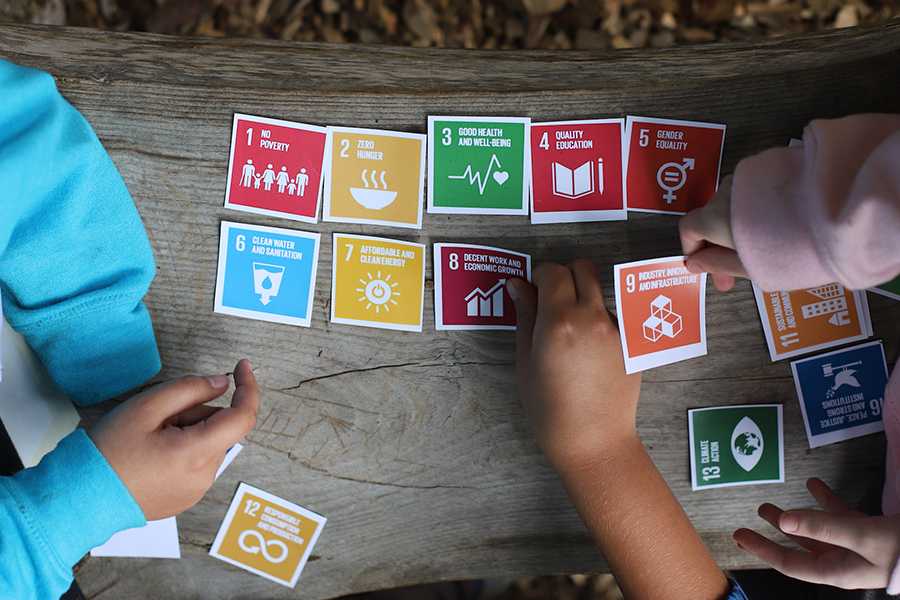 Colorful cards depicting the UN Sustainable Development are spread out on an outdoor bench as the hands of three young people reach to move the cards